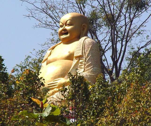 Large golden laughing Buddha statue surrounded by trees in Thailand